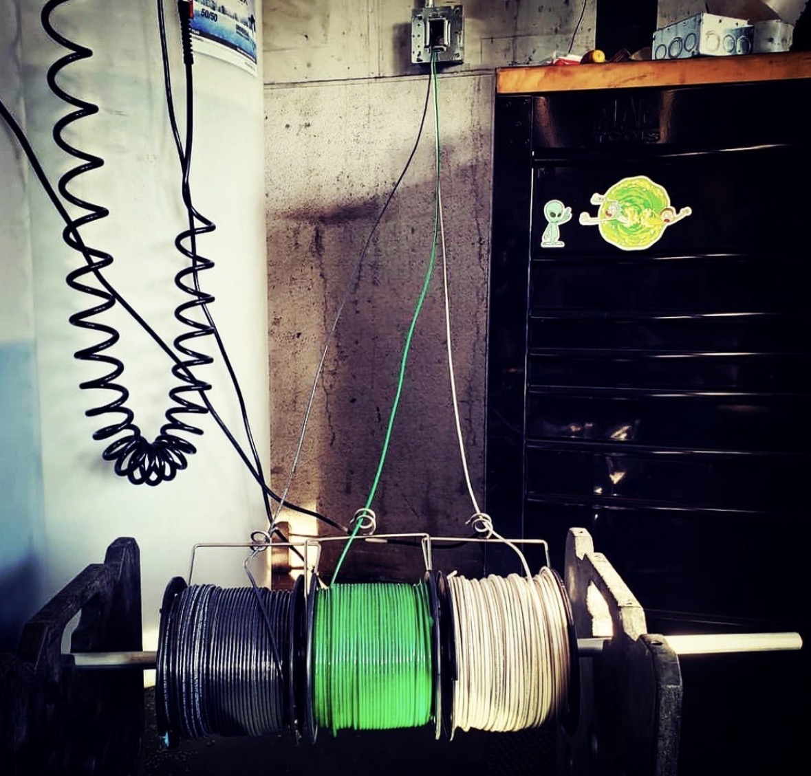 SpoolPig holding three wire spools (black, green, white) suspended from a workbench in an electrician's shop
